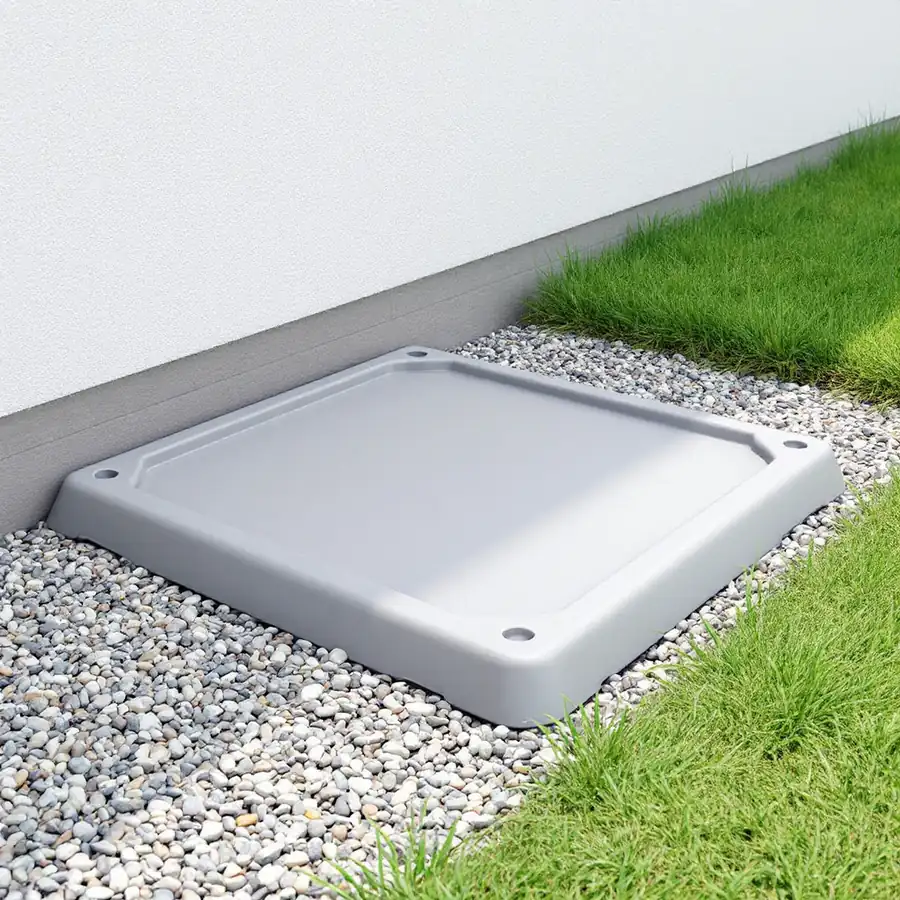 An empty grey moulded equipment pad lying level on a bed of gravel beside a white house wall with grass at the edge